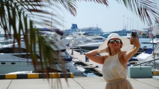Γυναίκα με καπέλο και γυαλιά ηλίου για το φόντο κάνει selfie στον κόλπο με γιοτ. Κορίτσι σε μια φούστα-ο ήλιος είναι κλώση και πανηγυρισμούς. 4 k αργή κίνηση. — Αρχείο Βίντεο