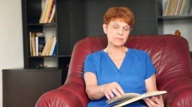 Yaşlı bir deri kanat sandalyede oturan kadın bir kitap okuma. 4k, ağır çekim