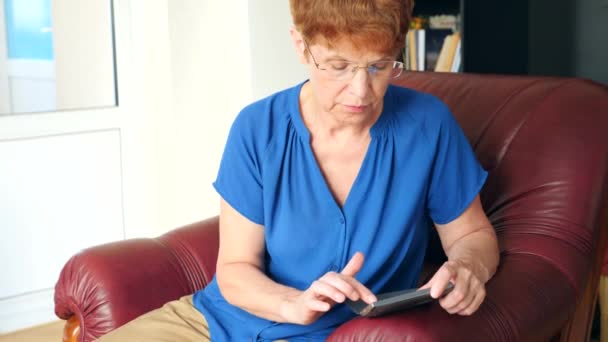 Vecchia signora seduta su una sedia ala in pelle, compone un messaggio sul suo smartphone. 4k, rallentatore — Video Stock