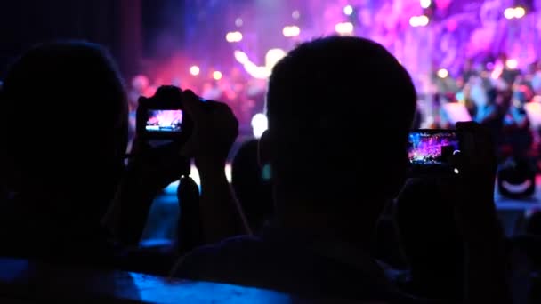 Kijkers schieten video op smartphones tijdens een concert van een rockband met een symfonieorkest. 4 k. Slowmotion — Stockvideo