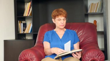 Yaşlı bir deri kanat sandalyede oturan kadın bir kitap okuma. 4k, ağır çekim