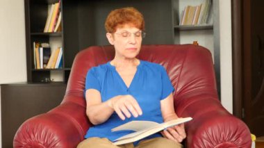 Yaşlı bir deri kanat sandalyede oturan kadın bir kitap okuma. 4k, ağır çekim