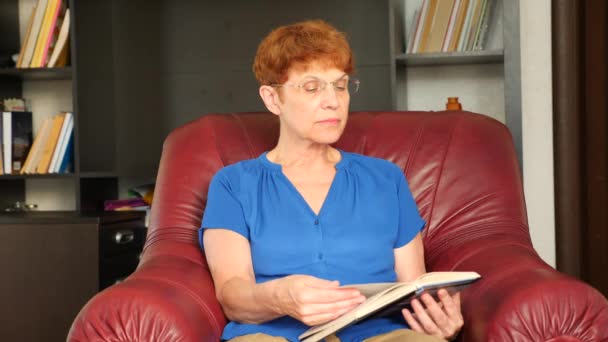 Alte Dame sitzt in einem ledernen Ohrensessel und liest ein Buch. 4k, Zeitlupe — Stockvideo