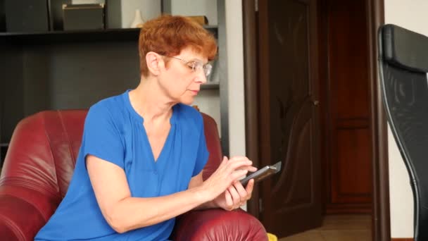 Starsza pani siedzi na krześle skrzydło skórzany, wybiera wiadomości w swoim smartfonie. 4k, zwolnionym tempie — Wideo stockowe