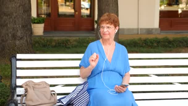 Una dulce anciana está sentada en un banco en un parque de la ciudad. Ella escucha música en auriculares desde el teléfono inteligente. Ella está bailando. 4. En cámara lenta. — Vídeos de Stock