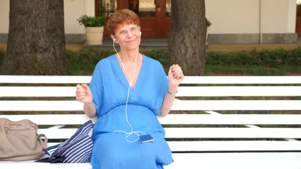 A sweet elderly lady is sitting on a bench in a city park. She listens to music on headphones from the smartphone. She is dancing. 4 . Slow-motion — Stock Video