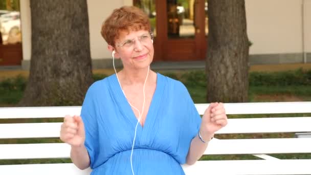 甜蜜的老太太正坐在一个城市公园的长椅上。她从智能手机听音乐头戴式耳机。她跳舞。4。慢动作 — 图库视频影像