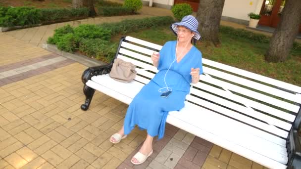 Słodki starsza pani siedzi na ławce w parku miejskim. Słucha muzyki na słuchawkach z smartphone. Ona tańczy. 4. Zwolnionym tempie — Wideo stockowe