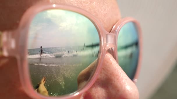 Cara de cerca de una mujer feliz con gafas de sol. La playa con la gente descansando se refleja en las gafas. 4k, cámara lenta — Vídeos de Stock