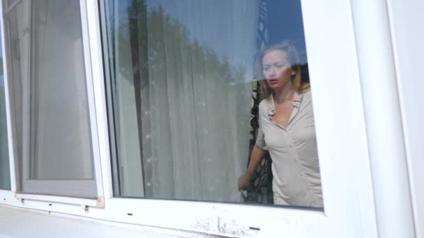 Una joven mujer parece asustada por detrás de las cortinas en la ventana, ella está asustada por el incidente que ocurrió en la calle. 4k, cámara lenta — Vídeo de stock