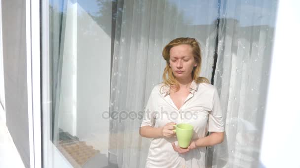Uma jovem atraente abre as cortinas em câmera lenta, olha para o nascer do sol, em pé junto à janela em sua casa. Com uma caneca de chá ou café. 4K. Movimento lento . — Vídeo de Stock