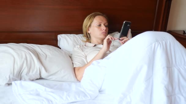 La donna è sdraiata a letto e utilizza uno smartphone. 4k, rallentatore — Video Stock