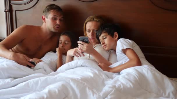 Una familia feliz está mirando algo en su teléfono inteligente mientras está acostado en la cama. 4k, cámara lenta . — Vídeo de stock