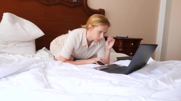 Estilo de vida autónomo. La mujer está trabajando detrás de un portátil en su cama en casa. 4k, cámara lenta — Vídeo de stock