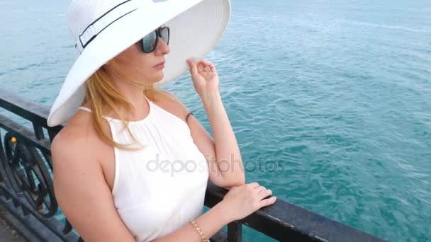 Uma mulher elegante com um chapéu branco de abas largas e óculos de sol desfruta da vista para o mar. 4k, câmera lenta — Vídeo de Stock