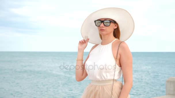 Una mujer elegante con un sombrero blanco de ala ancha y gafas de sol disfruta de la vista al mar. 4k, cámara lenta — Vídeo de stock