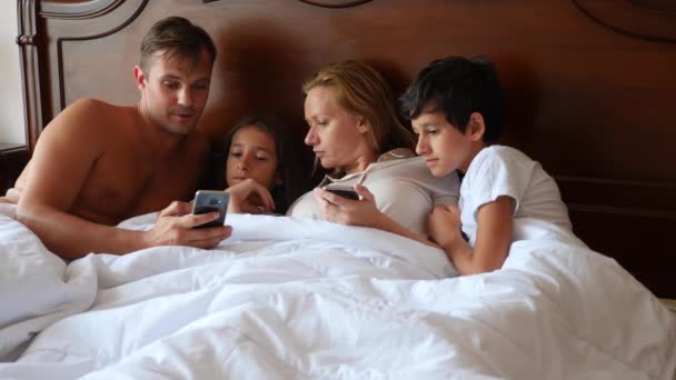 Szczęśliwa rodzina patrzy na coś na ich smartphone leżąc w łóżku. 4k, zwolnionym tempie. — Wideo stockowe