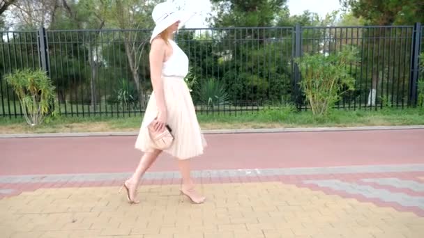 Una mujer elegante con un sombrero y un vestido de aire blanco pasea a lo largo del paseo marítimo. Disparos generales. 4k, cámara lenta — Vídeo de stock