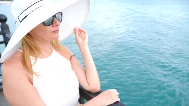 Uma mulher elegante com um chapéu branco de abas largas e óculos de sol desfruta da vista para o mar. 4k, câmera lenta — Vídeo de Stock