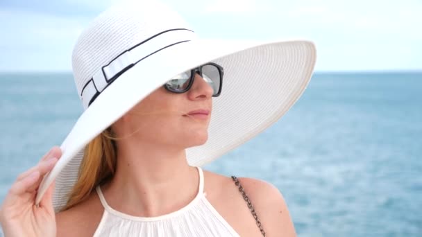 Μια κομψή γυναίκα με ένα άσπρο καπέλο με ευρύ γείσο και γυαλιά ηλίου απολαμβάνει τη θέα στη θάλασσα. 4k, αργή κίνηση — Αρχείο Βίντεο