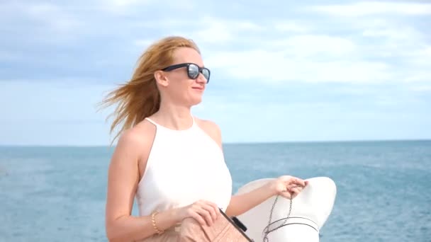 Елегантна жінка в білому широкоекранному капелюсі і сонцезахисних окулярах насолоджується видом на море. 4k, повільний рух — стокове відео