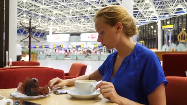 Mujer utiliza un teléfono inteligente en la sala de espera del aeropuerto. Ella bebe café. Expectativas de vuelo en el aeropuerto. 4k, cámara lenta — Vídeo de stock