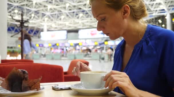 Mulher usa um smartphone na sala de espera do aeroporto. Ela bebe café. Expectativas de voo no aeroporto. 4k, câmera lenta — Vídeo de Stock