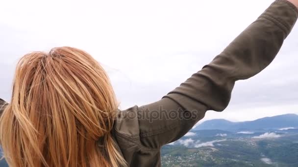 Молодая блондинка-путешественница на вершине горы поднимает руки вверх. ветер развивает ее волосы. 4k. Медленное движение — стоковое видео