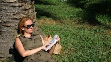 sarışın kadın parkta çim bir ağacın altında oturup kitap okuma. 4k, ağır çekim