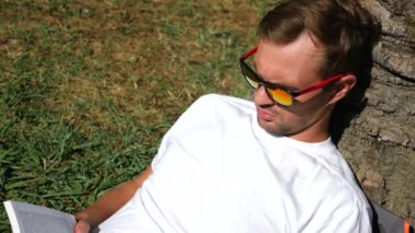 Yakışıklı adam parkta bir ağacın altında çimlerin üzerine oturmuş kitap, 4 k, yavaş hareket okuma