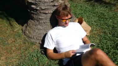 Yakışıklı adam parkta bir ağacın altında çimlerin üzerine oturmuş kitap, 4 k, yavaş hareket okuma