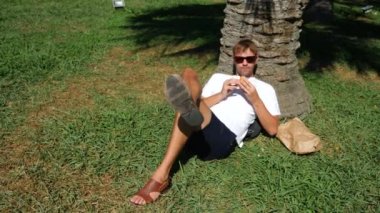 Çekici adam çim Park'ta bir ağacın altında oturup taze hamburger yeme. 4k, ağır çekim