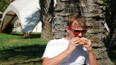 Çekici adam çim Park'ta bir ağacın altında oturup taze hamburger yeme. 4k, ağır çekim