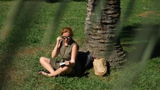 Blonde Frau liest das Buch im Park, sitzt auf dem Gras, unter einem Baum. 4k, Zeitlupe — Stockvideo