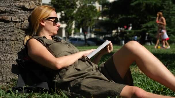 Blonde Frau liest das Buch im Park, sitzt auf dem Gras, unter einem Baum. 4k, Zeitlupe — Stockvideo