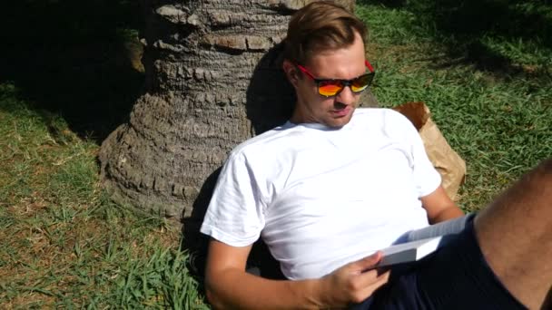 Schöner Mann sitzt auf dem Gras im Park unter einem Baum, liest Buch, 4k, Zeitlupe — Stockvideo