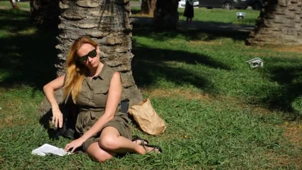 Femme blonde lisant le livre dans le parc, assise sur l'herbe, sous un arbre. 4k, au ralenti — Video