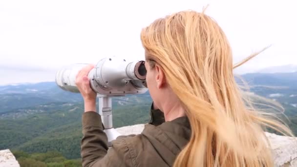 Блондинка-путешественница смотрит в телескоп на вершине горы. 4k, slow-motion — стоковое видео