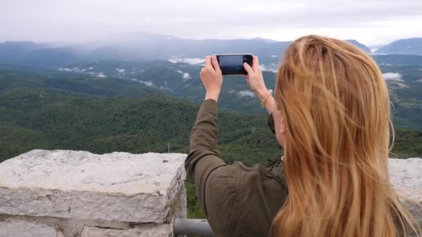 Mujer joven en la cima de la montaña tomando una selfie en cámara lenta. Joven excursionista tomando fotografías de una vista de las montañas desde la cima de la montaña hasta su teléfono. 4k — Vídeos de Stock