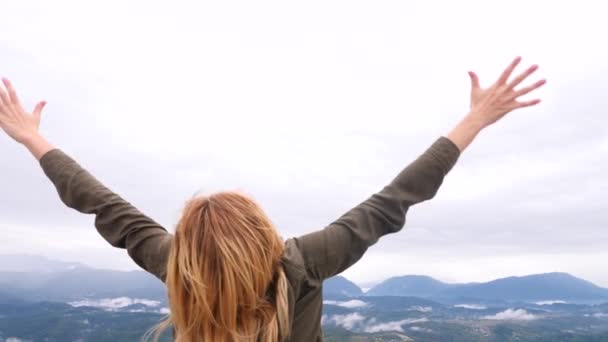 Молодая блондинка-путешественница на вершине горы поднимает руки вверх. ветер развивает ее волосы. 4k. Медленное движение — стоковое видео