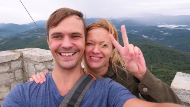 Pareja feliz hombre y mujer son fotografiados en la cima de una montaña en el fondo de las montañas. el viento desarrolla el cabello. 4k, cámara lenta — Vídeo de stock