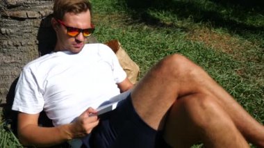 Yakışıklı adam parkta bir ağacın altında çimlerin üzerine oturmuş kitap, 4 k, yavaş hareket okuma