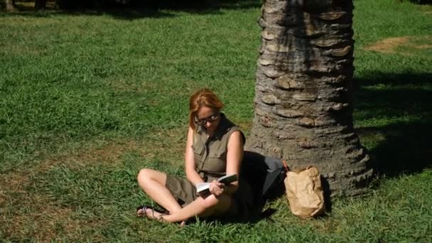 Ξανθιά γυναίκα διαβάζοντας το βιβλίο στο πάρκο, συνεδρίαση για το γρασίδι, κάτω από ένα δέντρο. 4k, αργή κίνηση — Αρχείο Βίντεο