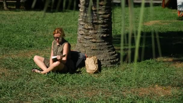 Blonde vrouw lezen van het boek in het park, zittend op het gras, onder een boom. 4k, slow-motion — Stockvideo