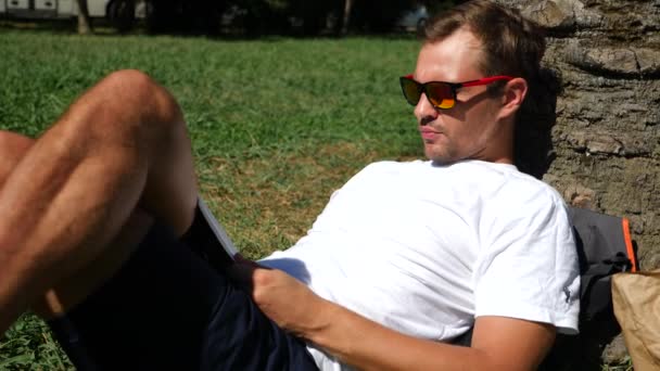 Yakışıklı adam parkta bir ağacın altında çimlerin üzerine oturmuş kitap, 4 k, yavaş hareket okuma — Stok video