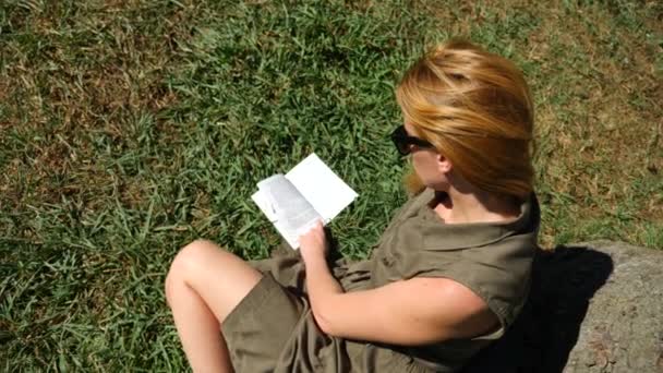 Blonde vrouw lezen van het boek in het park, zittend op het gras, onder een boom. 4k, slow-motion — Stockvideo