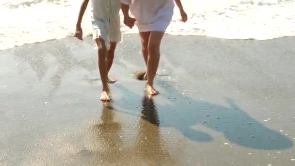 Mutter und Tochter gehen am Ufer des Meeres spazieren, Wasserspritzer auf den Füßen, langsame Bewegungen. die Welle schließt die Beine derjenigen, die gehen. 4k — Stockvideo