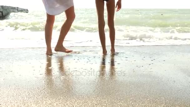 Μητέρα και κόρη με τα πόδια στην παραλία, πιτσιλιές του νερού στα πόδια τους, αργές κινήσεις. Το κύμα κλείνει τα πόδια εκείνων που περπατούν. 4k — Αρχείο Βίντεο