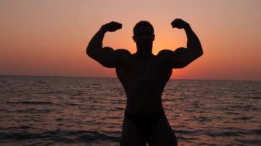 bir erkek vücut geliştirmeci silüeti. gün batımında sahilde poz güzel atlet. 4 k. yavaş çekim