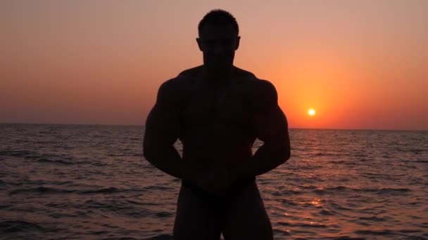 Sziluettjét egy férfi testépítő. gyönyörű sportoló pózol a tengerparton naplementekor. 4 k., lassú lövés — Stock videók